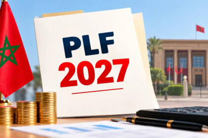 Un document portant la mention « PLF 2027 » posé à côté d’un drapeau marocain, de piles de pièces dorées, d’une calculatrice et d’un stylo, devant un bâtiment officiel.