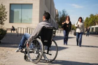 Homme en fauteuil roulant se déplaçant sur un parvis urbain pavé, entouré de passants et de bâtiments modernes, symbolisant l'inclusion sociale et les dispositifs d'aide au logement.