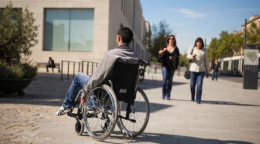 Homme en fauteuil roulant se déplaçant sur un parvis urbain pavé, entouré de passants et de bâtiments modernes, symbolisant l'inclusion sociale et les dispositifs d'aide au logement.