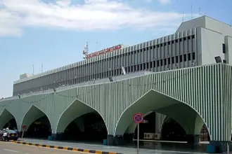 Façade verte à rayures et à grandes arcades du terminal de l’aéroport de Tripoli, avec des véhicules garés devant.