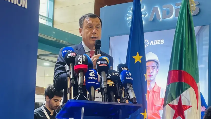 Un homme en costume sombre s'exprime au micro derrière un pupitre aux couleurs de l'Union européenne, entouré de micros de médias, devant les drapeaux européens et algériens.