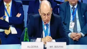 Un homme en costume sombre et lunettes lit un texte devant un micro, assis derrière les plaques « Algeria » et « Mr. Bourai », avec deux participants assis à ses côtés.