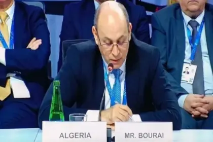 Un homme en costume sombre et lunettes lit un texte devant un micro, assis derrière les plaques « Algeria » et « Mr. Bourai », avec deux participants assis à ses côtés.