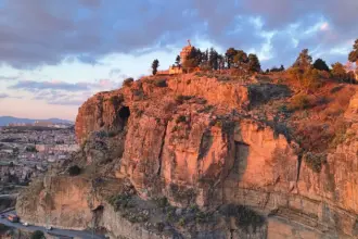 Falaise rocheuse éclairée par une lumière rasante, surmontée d’un monument entouré d’arbres. En contrebas, une route en corniche où circule un bus orange, et la ville sous un ciel nuageux rose et bleu.