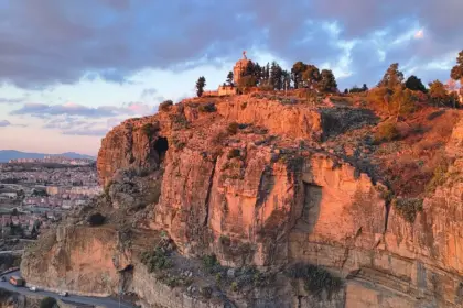 Falaise rocheuse éclairée par une lumière rasante, surmontée d’un monument entouré d’arbres. En contrebas, une route en corniche où circule un bus orange, et la ville sous un ciel nuageux rose et bleu.