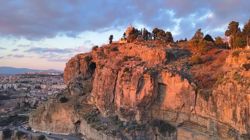 Falaise rocheuse éclairée par une lumière rasante, surmontée d’un monument entouré d’arbres. En contrebas, une route en corniche où circule un bus orange, et la ville sous un ciel nuageux rose et bleu.