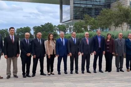 Treize personnes, dix hommes et trois femmes en tenue de ville, posent côte à côte en ligne devant un bâtiment moderne à façade vitrée, sur une esplanade pavée bordée d'arbres, sous un ciel nuageux.