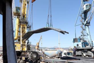 Grues portuaires soulevant un lot de barres d’acier sur un quai du port de Béjaïa, avec camions et ouvriers au second plan