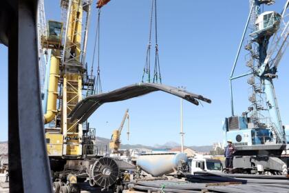 Grues portuaires soulevant un lot de barres d’acier sur un quai du port de Béjaïa, avec camions et ouvriers au second plan