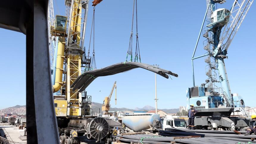 Grues portuaires soulevant un lot de barres d’acier sur un quai du port de Béjaïa, avec camions et ouvriers au second plan