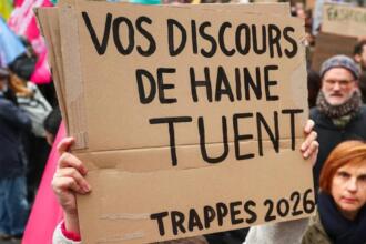 Une main tient une pancarte en carton où l’on lit en lettres noires « Vos discours de haine tuent – Trappes 2026 », devant des manifestants et des drapeaux colorés.