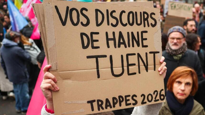 Une main tient une pancarte en carton où l’on lit en lettres noires « Vos discours de haine tuent – Trappes 2026 », devant des manifestants et des drapeaux colorés.