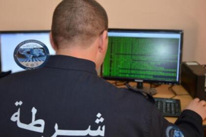 Policier en uniforme vu de dos, assis devant deux écrans d’ordinateur dont l’un affiche des lignes de code vert sur fond noir, avec le mot « شرطة » (police) inscrit sur son dos.