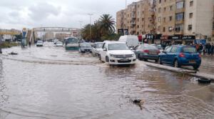 Voitures et minibus roulant dans une rue inondée devant des immeubles, un SUV blanc soulevant l’eau au premier plan