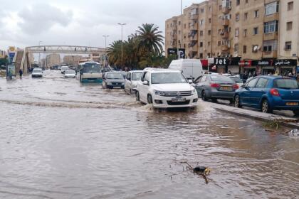 Voitures et minibus roulant dans une rue inondée devant des immeubles, un SUV blanc soulevant l’eau au premier plan