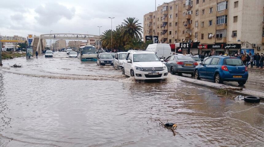 Voitures et minibus roulant dans une rue inondée devant des immeubles, un SUV blanc soulevant l’eau au premier plan
