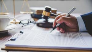 Main d'un juriste signant un contrat posé sur un porte-bloc, avec un maillet de juge et une balance de la justice en arrière-plan