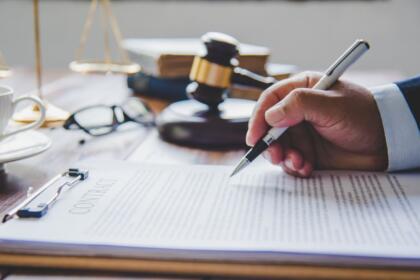 Main d'un juriste signant un contrat posé sur un porte-bloc, avec un maillet de juge et une balance de la justice en arrière-plan