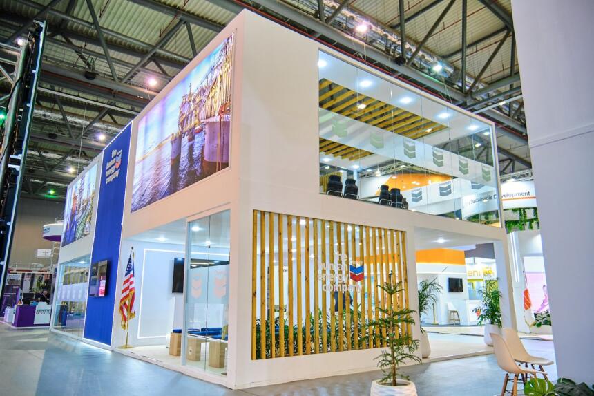 Stand de Chevron au NAPEC 2025, à Oran : structure à deux niveaux avec salle de réunion vitrée à l'étage, grand panneau lumineux représentant une plateforme offshore et drapeau américain à l'entrée.