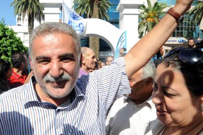 Zied El-Heni, cheveux gris et moustache, sourit en levant le bras en signe de victoire au milieu d’une foule, devant un portail blanc orné d’un drapeau.